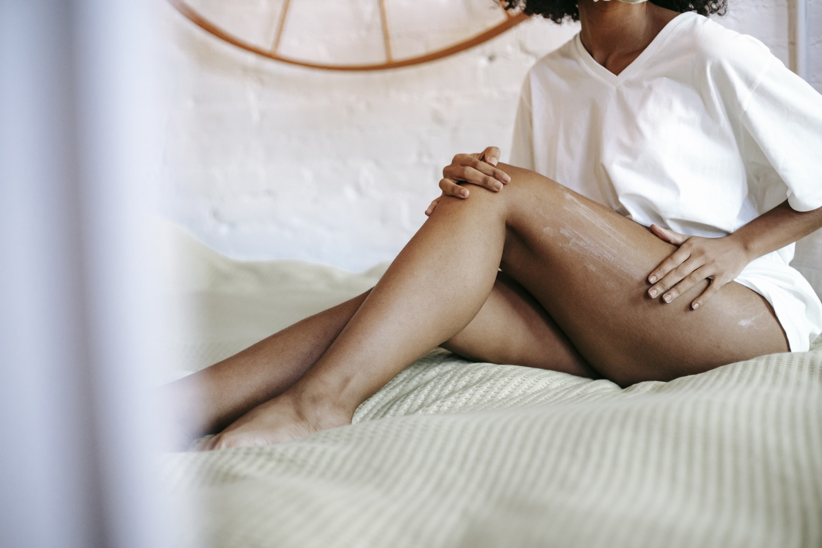 Side view of unrecognizable female in white t shirt sitting on bed and applying lotion on leg while resting in weekend
